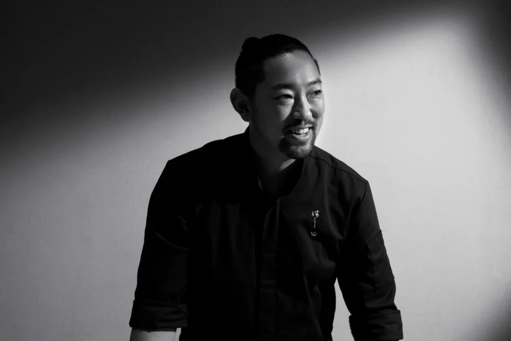 A black-and-white portrait of a smiling man, Chef Kenjiro "Hatch" Hashida, wearing a dark chef's jacket with small Japanese embroidery. Dramatic spotlighting illuminates his face and creates a sharp, diagonal shadow across the background behind him.
