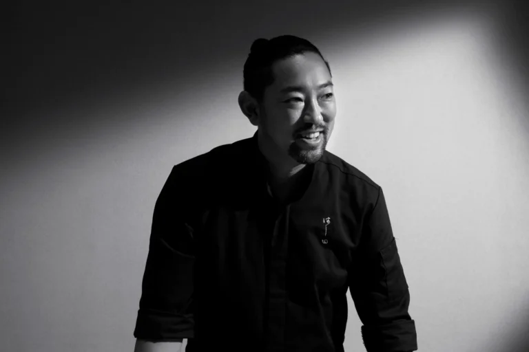 A black-and-white portrait of a smiling man, Chef Kenjiro "Hatch" Hashida, wearing a dark chef's jacket with small Japanese embroidery. Dramatic spotlighting illuminates his face and creates a sharp, diagonal shadow across the background behind him.