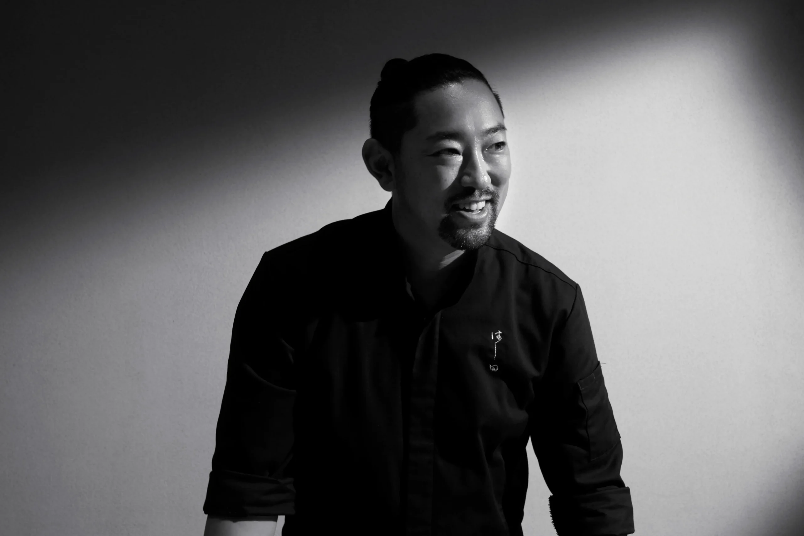 A black-and-white portrait of a smiling man, Chef Kenjiro "Hatch" Hashida, wearing a dark chef's jacket with small Japanese embroidery. Dramatic spotlighting illuminates his face and creates a sharp, diagonal shadow across the background behind him.