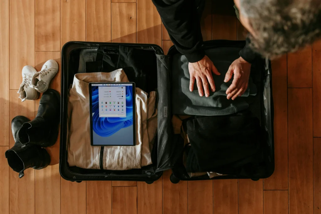 An overhead shot shows a man packing dark clothing into an open black suitcase resting on a wooden floor. A Microsoft Surface tablet displaying the Windows start menu sits on top of folded clothes inside the luggage, next to pairs of shoes scattered nearby.