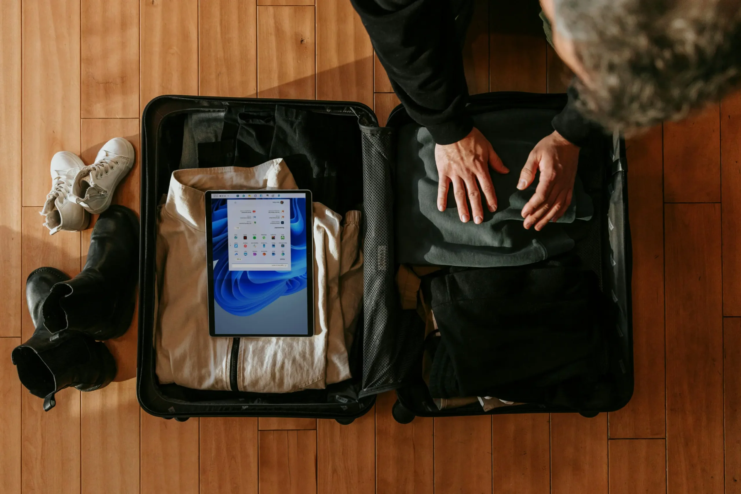 An overhead shot shows a man packing dark clothing into an open black suitcase resting on a wooden floor. A Microsoft Surface tablet displaying the Windows start menu sits on top of folded clothes inside the luggage, next to pairs of shoes scattered nearby.