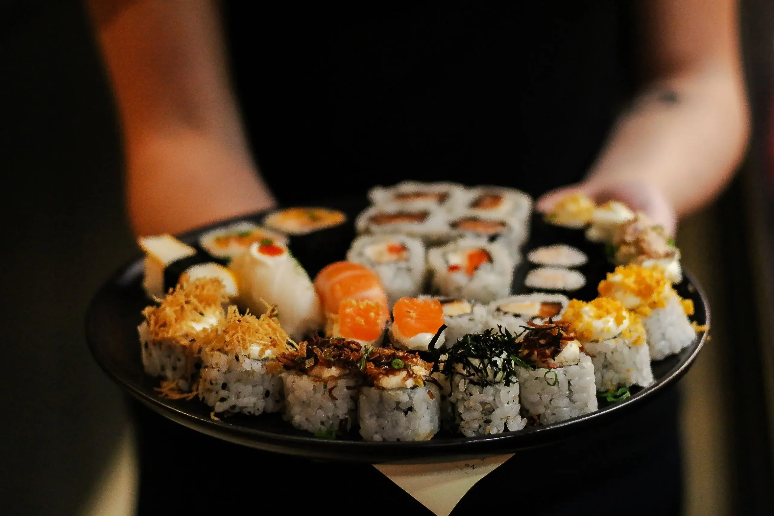 A person holds out a large round black platter filled with a colorful variety of sushi rolls and nigiri. The selection includes rolls topped with salmon, crispy toppings, seaweed flakes, and creamy sauces.