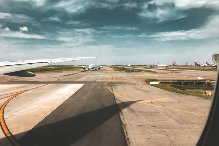 A view from inside an airplane window shows a wing extending over a wide airport tarmac. Another commercial airliner is positioned further down the taxiway under a dramatic, cloudy sky.