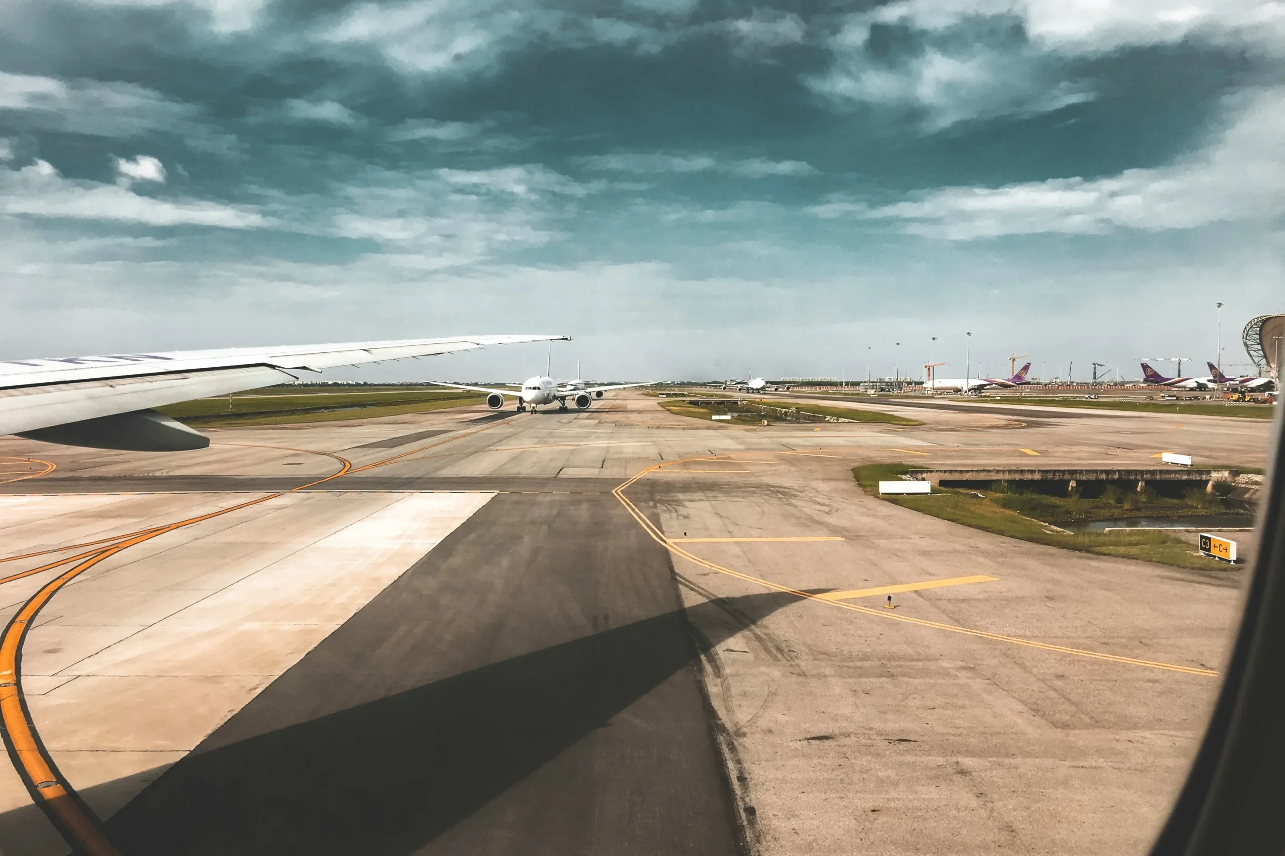 A view from inside an airplane window shows a wing extending over a wide airport tarmac. Another commercial airliner is positioned further down the taxiway under a dramatic, cloudy sky.
