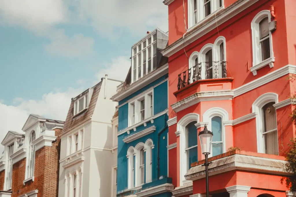The image shows a row of elegant, multi-story terraced houses painted in vibrant colors, featuring a striking coral red building next to a bright blue one. A classic black street lamp stands in the foreground, highlighting the architecture's ornate white trim, arched windows, and wrought-iron balconies against a partly cloudy sky.