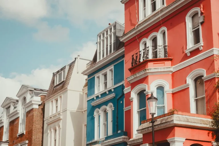The image shows a row of elegant, multi-story terraced houses painted in vibrant colors, featuring a striking coral red building next to a bright blue one. A classic black street lamp stands in the foreground, highlighting the architecture's ornate white trim, arched windows, and wrought-iron balconies against a partly cloudy sky.