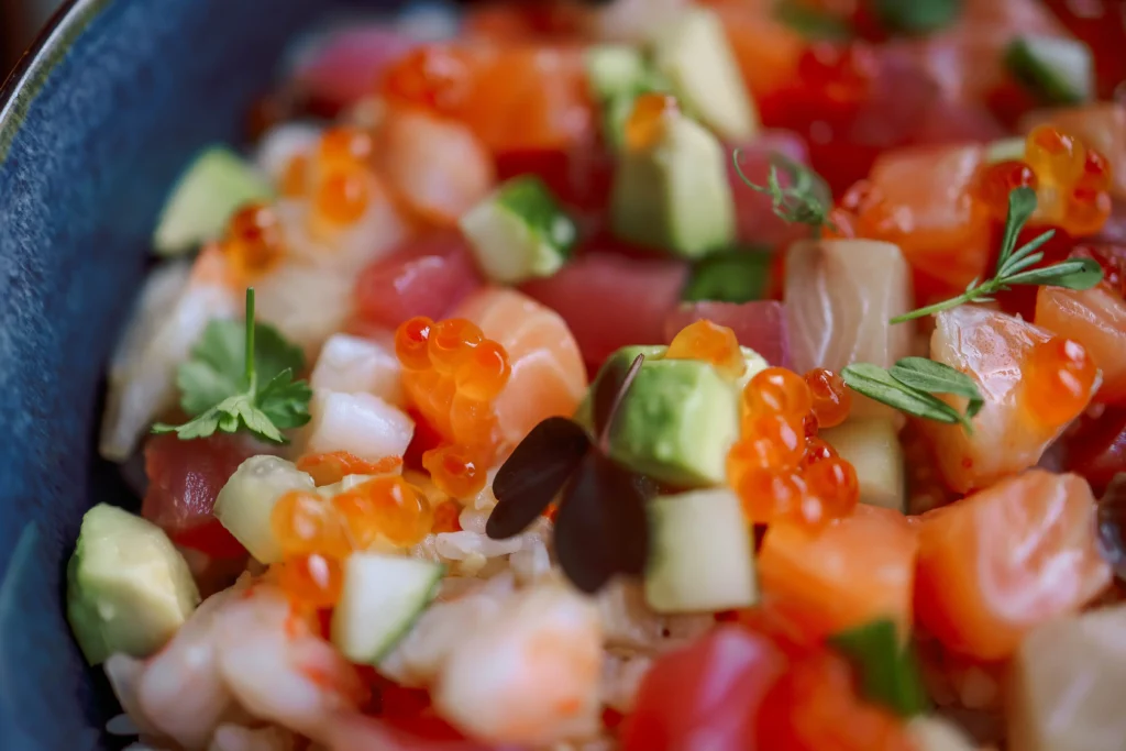 A close-up view of a colorful poke bowl filled with cubed raw salmon, tuna, avocado, cucumber, and shiny orange fish roe. Small green herb garnishes and microgreens are scattered across the top for added texture and detail.