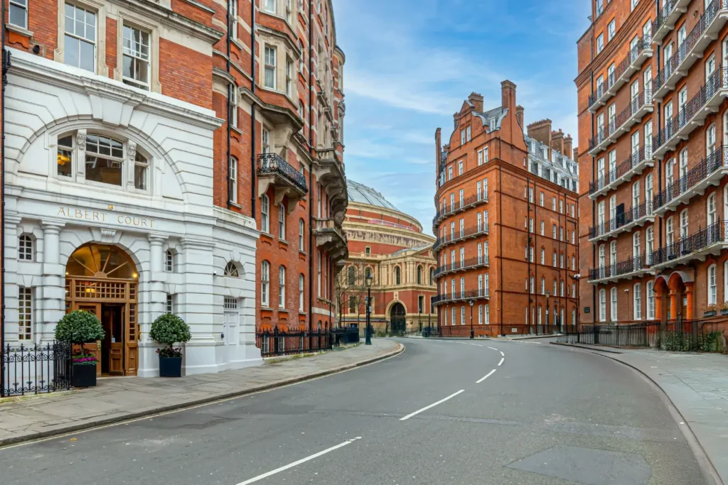 A street view in London looking down a curved asphalt road lined with grand, multi-story red brick residential buildings, including the prominent white-arched entrance of Albert Court on the left. In the background, the distinctive domed structure of the Royal Albert Hall is visible under a bright, partly cloudy sky.