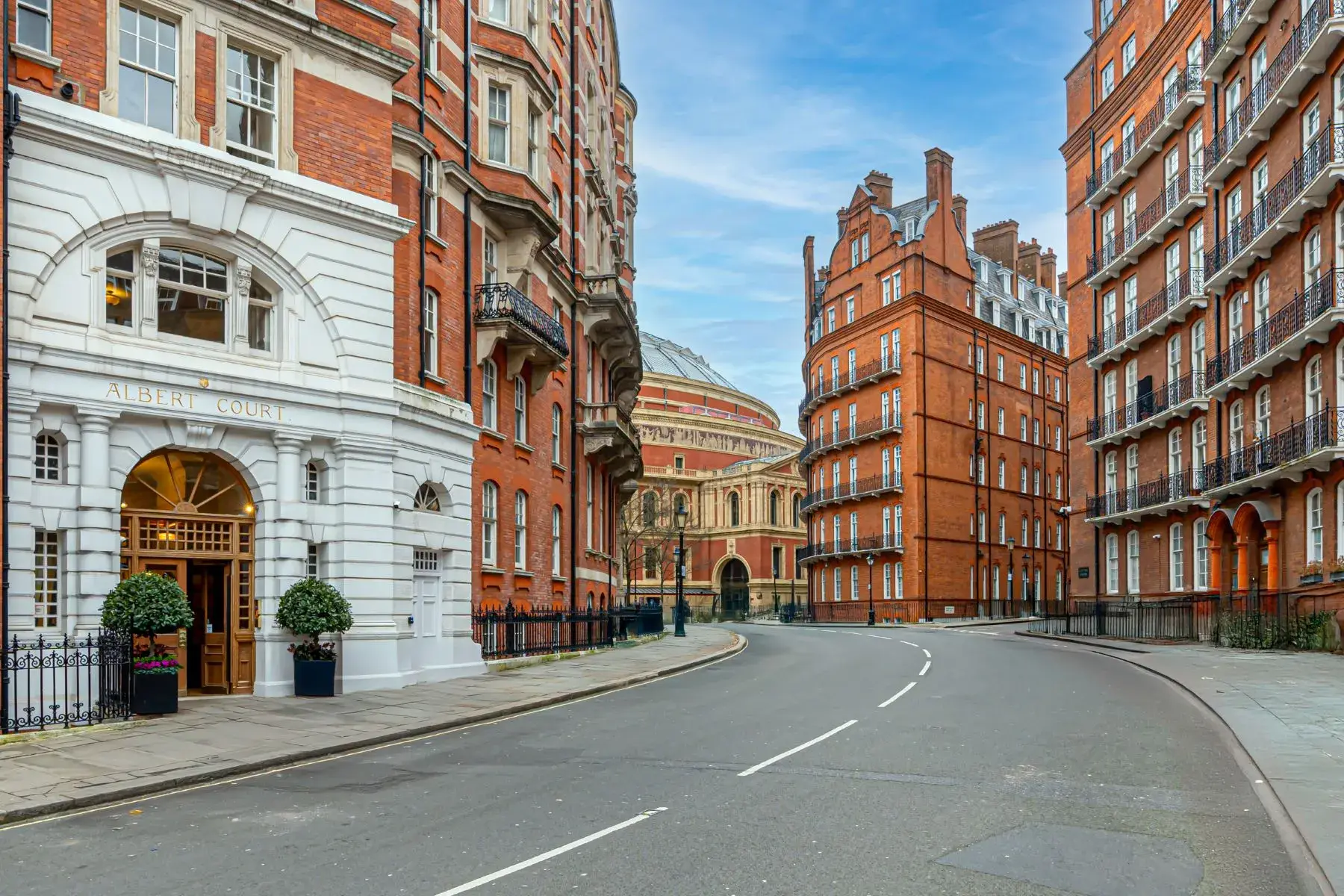 A street view in London looking down a curved asphalt road lined with grand, multi-story red brick residential buildings, including the prominent white-arched entrance of Albert Court on the left. In the background, the distinctive domed structure of the Royal Albert Hall is visible under a bright, partly cloudy sky.