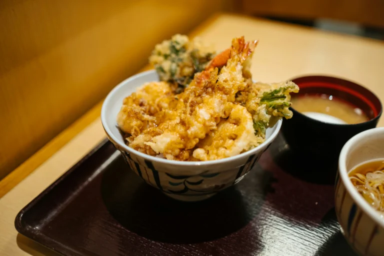 A bowl of golden, crispy tendon (tempura rice bowl) sits on a dark wooden tray alongside small bowls of miso soup and noodles. The lightly drizzled tempura pieces, including shrimp and vegetables, are piled high over rice in a patterned ceramic bowl.