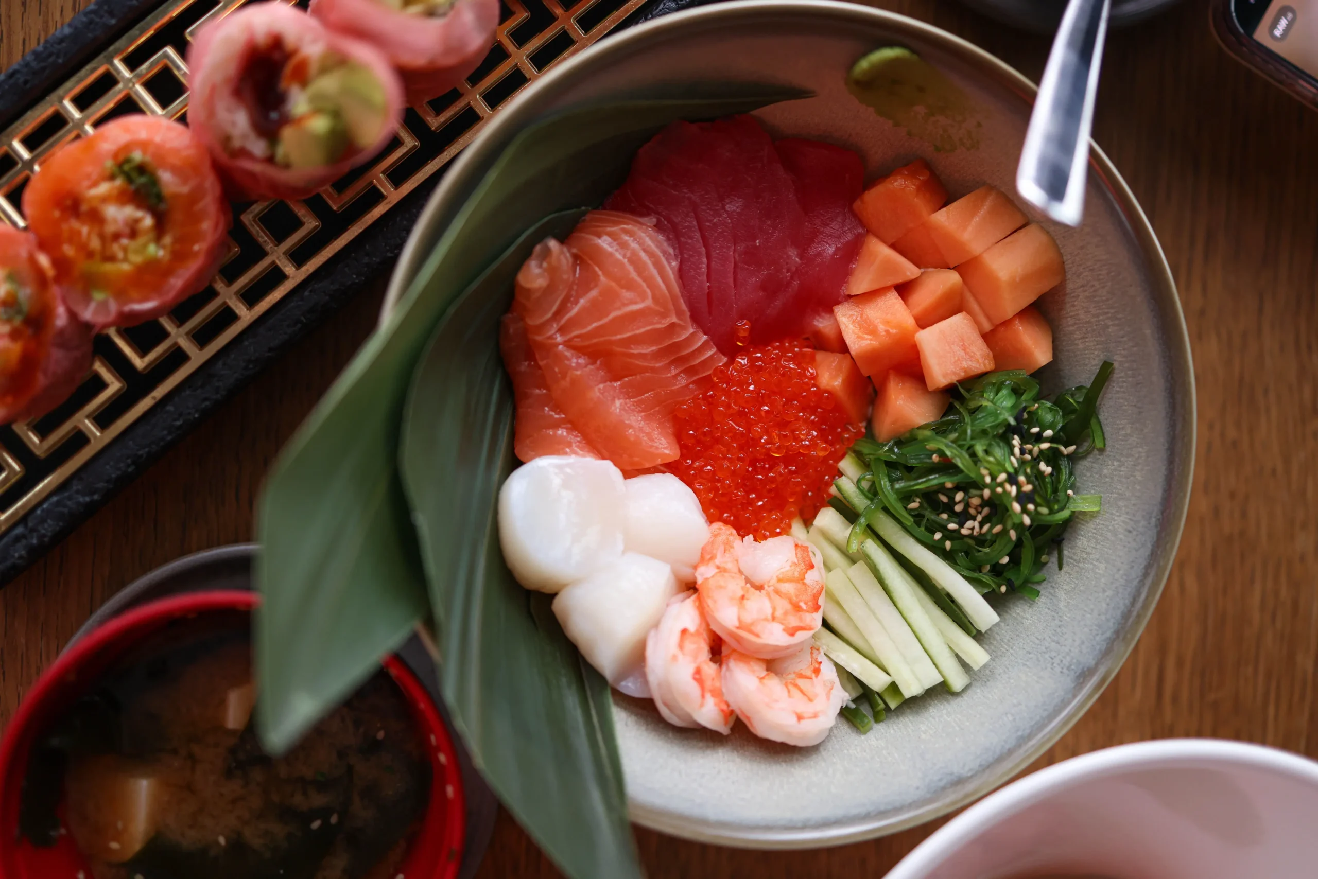 An overhead view features a colorful Japanese seafood bowl filled with sliced salmon, tuna, raw scallops, cooked shrimp, salmon roe, seaweed salad, and cucumber. The dish is set on a wooden dining table next to a row of sushi rolls and a bowl of miso soup.