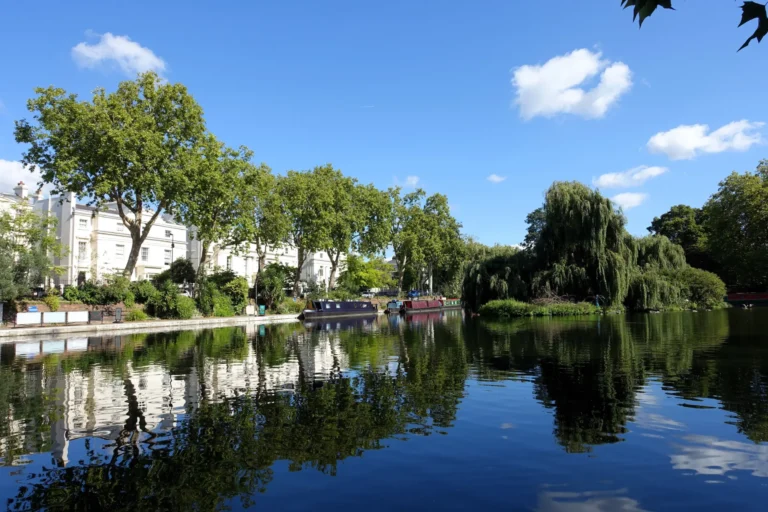 A scenic view of a calm canal shows narrowboats moored alongside lush green trees and elegant white buildings. The clear blue sky and surrounding greenery are beautifully reflected on the dark, still water's surface.