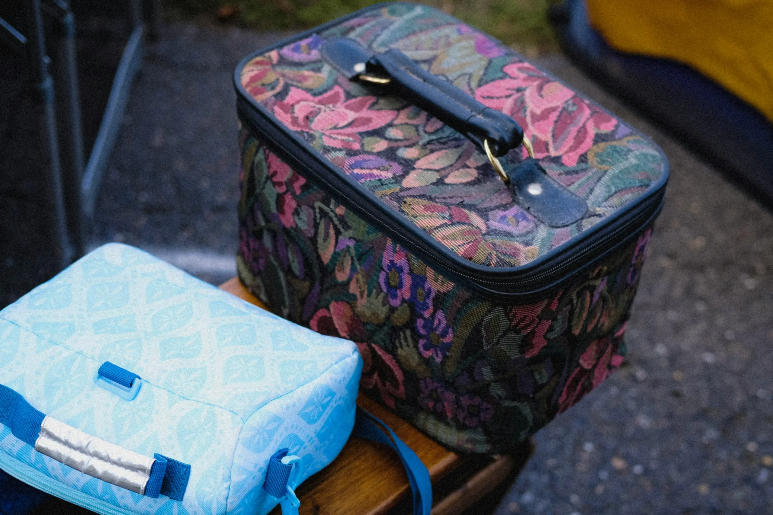 A close-up shot displays a vintage-style tapestry makeup case with a pink and purple floral pattern and a black handle sitting outdoors on a wooden surface. Next to it rests a modern light blue lunch bag or small cooler bag featuring a geometric white pattern and a carrying strap.