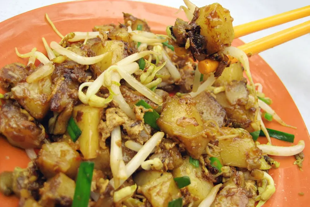 A close-up shot shows a plate of fried carrot cake, a popular Teochew dish featuring stir-fried radish cubes, bean sprouts, and green onions on an orange plate. A pair of yellow chopsticks lifts a piece of the savory, golden-brown radish cake from the pile.