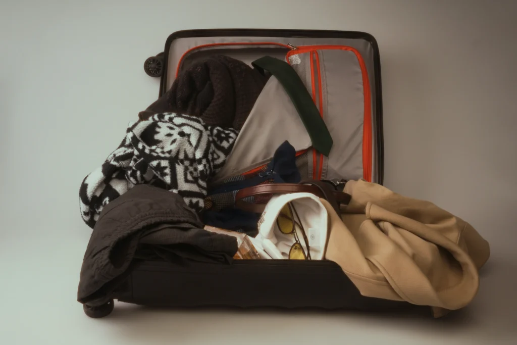 An open black rolling suitcase rests against a plain background, packed with various clothing items and travel accessories. The contents include a black-and-white patterned sweater, a beige jacket, folded dark trousers, sunglasses, and a leather belt.