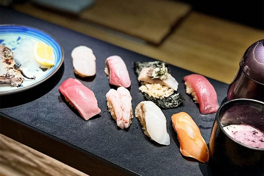 A selection of assorted sushi on a black serving platter, including tuna, salmon, white fish, shrimp, and seaweed-wrapped sushi, with a dipping sauce and lemon wedges on the side.