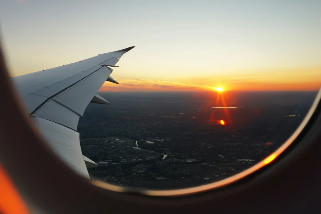 A view from an airplane window captures the wing extending over a sprawling landscape as the sun sets on the horizon. The sky glows with vibrant shades of orange and yellow, casting a warm light over the earth below.