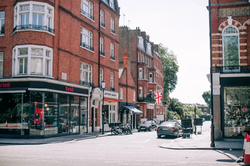 The image shows a sunny street corner in London featuring classic multi-story red brick buildings with storefronts on the ground floor. A Union Jack flag hangs from one of the buildings, while cars and motorcycles are parked along the side of the road.