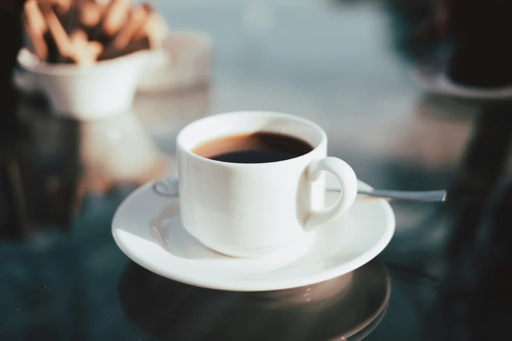 In the image, a white ceramic coffee cup filled with dark coffee rests on a matching saucer atop a reflective glass table. The background is softly blurred with a shallow depth of field, hinting at a cozy cafe setting.