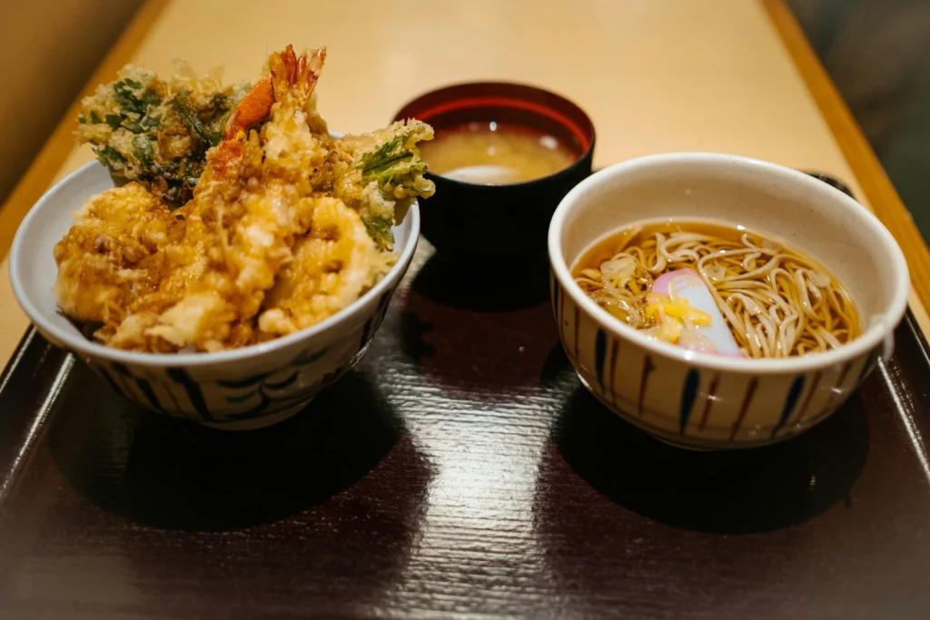 A traditional Japanese meal set is served on a dark tray atop a wooden table. The meal features a rice bowl topped with crispy golden tempura alongside a small bowl of hot noodle soup in a clear broth.