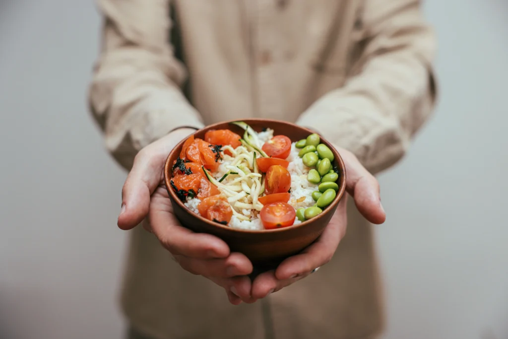 A person holding out a small wooden bowl filled with a fresh poke bowl over a bed of white rice. The dish is neatly arranged with bright toppings, including diced salmon, sliced cherry tomatoes, edamame, and shredded vegetables.