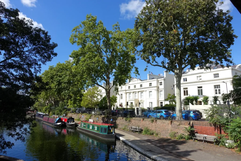 A scenic view of several colorful narrowboats moored along a calm canal, with a paved walkway and benches on the right bank. Tall green trees and elegant, white multi-story townhouse buildings stand under a bright blue sky with scattered clouds in the background.