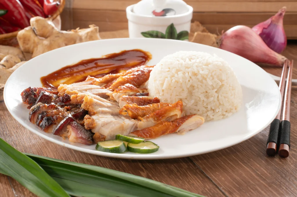 A large white plate holds a mound of white rice served alongside a selection of roasted meats, including crispy pork belly and roasted duck, drizzled with a dark savory sauce. Fresh cucumber slices rest on the plate, with wooden chopsticks, whole red onions, and ginger root visible in the background on a wooden table.