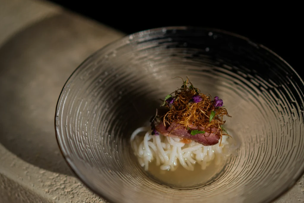 A gourmet dish is beautifully presented in a textured glass bowl, featuring a nest of translucent white noodles resting in a shallow, clear broth. The noodles are topped with a slice of seared meat, crispy golden-brown threads, and delicate purple edible flowers under dramatic, warm lighting.