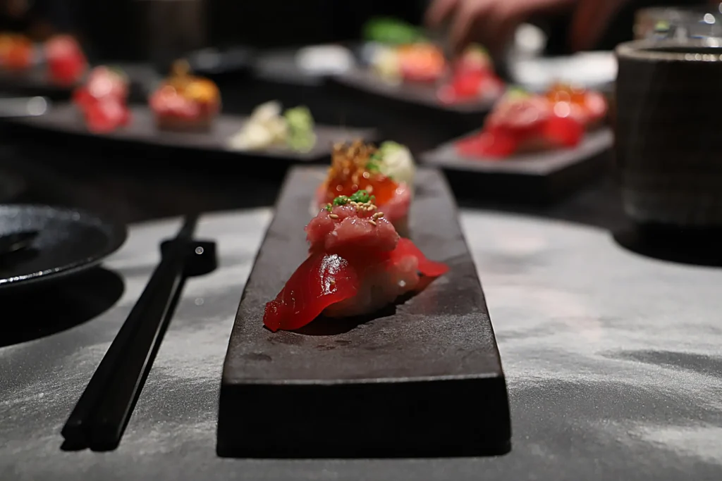 A close-up view shows gourmet tuna nigiri topped with minced tuna, sesame seeds, and microgreens served on a long, dark stone platter. Black chopsticks rest alongside the dish on a moody stone table, with additional blurred sushi platters setting the scene in the background.