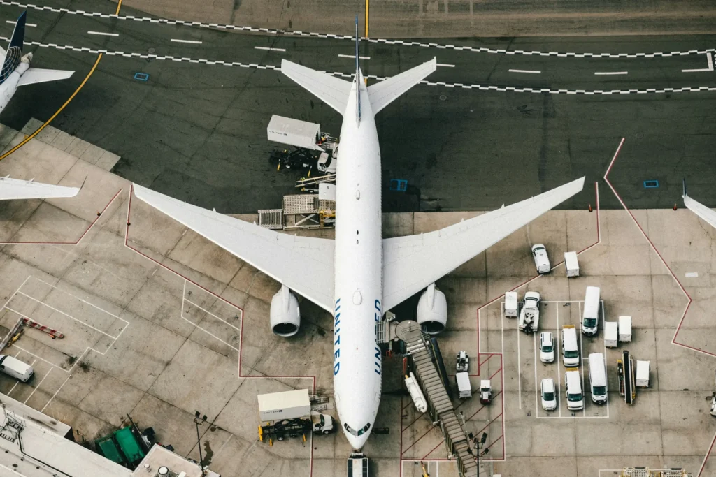 An overhead, top-down view shows a large white United Airlines passenger airplane parked at an airport terminal gate. Various ground service vehicles, including cargo loaders and white vans, are positioned around the aircraft on the tarmac.