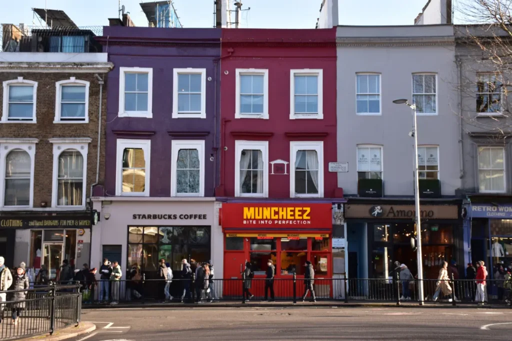A row of multi-colored, historic London-style buildings features storefronts along the street level, including a Starbucks Coffee and a bright red Muncheez restaurant. Pedestrians walk along the sidewalk in front of the shops under a clear, bright sky.