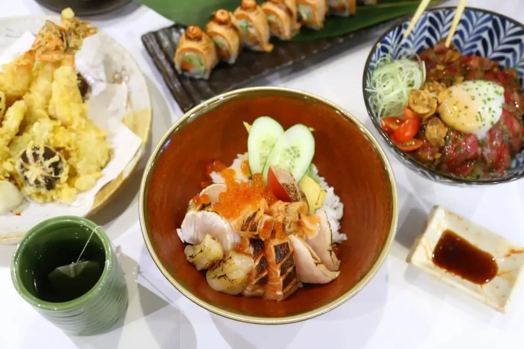 A large bowl of chirashi don topped with seared sashimi, salmon roe, and cucumber slices sits at the center of a table spread. Surrounding the main bowl are side dishes including tempura, sushi rolls, a beef rice bowl with a poached egg, and a cup of green tea.