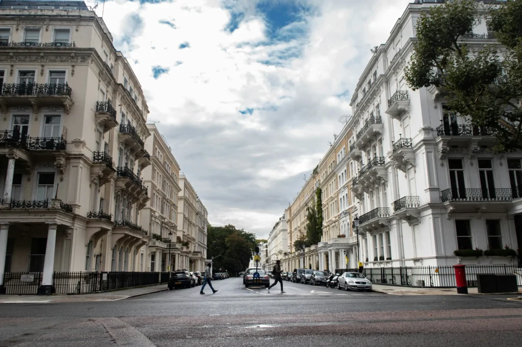 A ground-level view of a wide city street flanked by elegant, multi-story white and cream townhouse buildings with ornate balconies. Pedestrians cross the asphalt road under a bright sky filled with dramatic white and gray clouds.