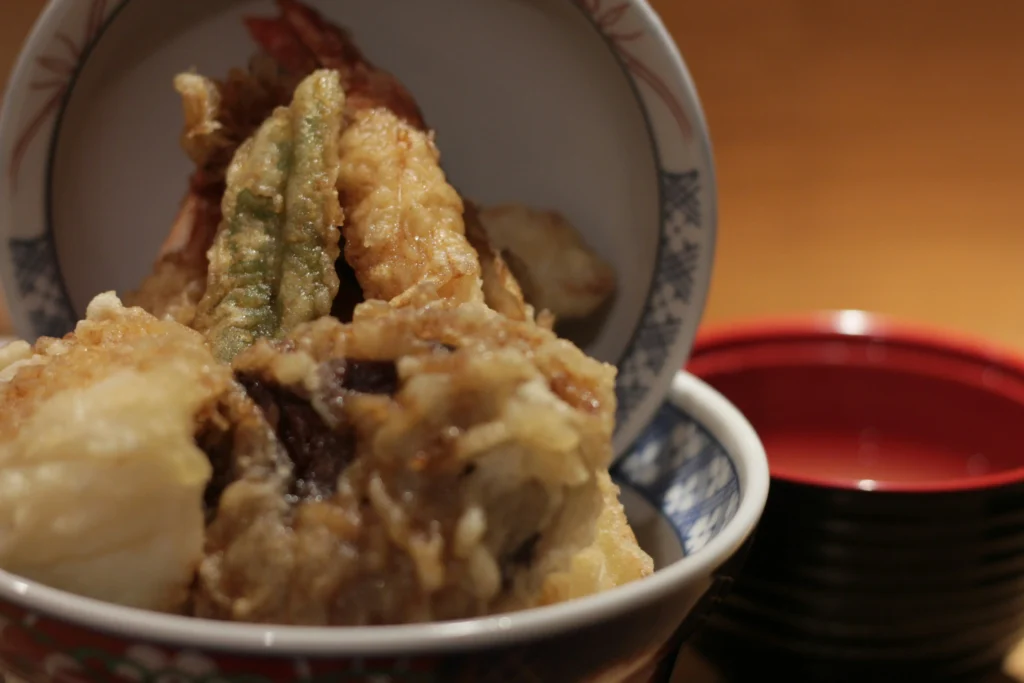 A close-up view of a Japanese tempura rice bowl (tendon) overflowing with crispy fried shrimp and vegetables, with the bowl's lid resting vertically behind the food. A small red and black lacquer bowl sits in the soft-focused background against a warm, wooden backdrop.
