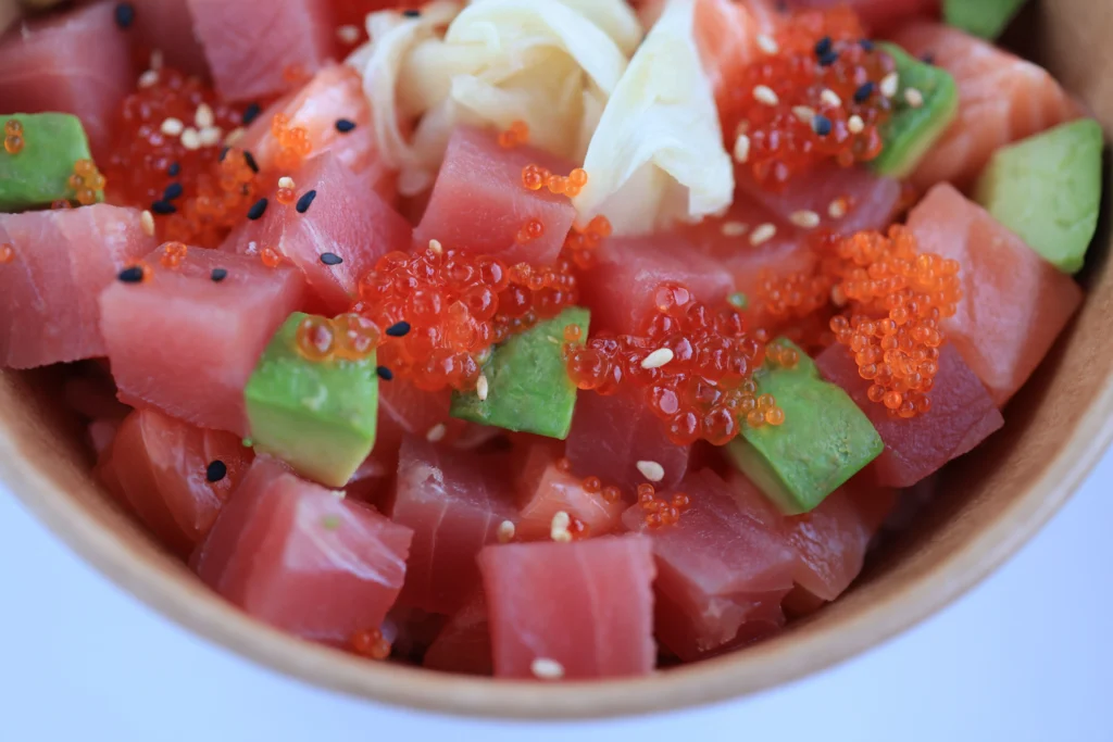 A close-up view of a colorful poke bowl filled with cubed raw tuna, salmon, and avocado. The dish is topped with glistening orange fish roe, pickled ginger, and a sprinkle of sesame seeds.