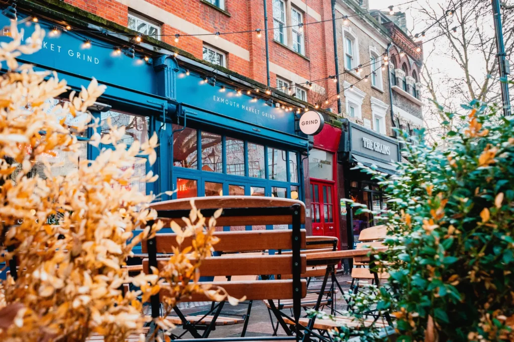 A street-level view of a charming café named "Exmouth Market Grind," featuring a vibrant blue storefront and string lights hung overhead. Wooden outdoor dining chairs are positioned in the foreground, framed by golden autumn leaves on the left and green foliage on the right.