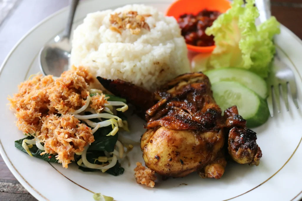 A plate of Indonesian-style food features a mound of white rice topped with fried shallots alongside pieces of dark, glazed grilled chicken. The dish is accompanied by fresh lettuce and sliced cucumbers, an orange cup of chili paste, and a side of vegetable salad topped with spiced grated coconut.