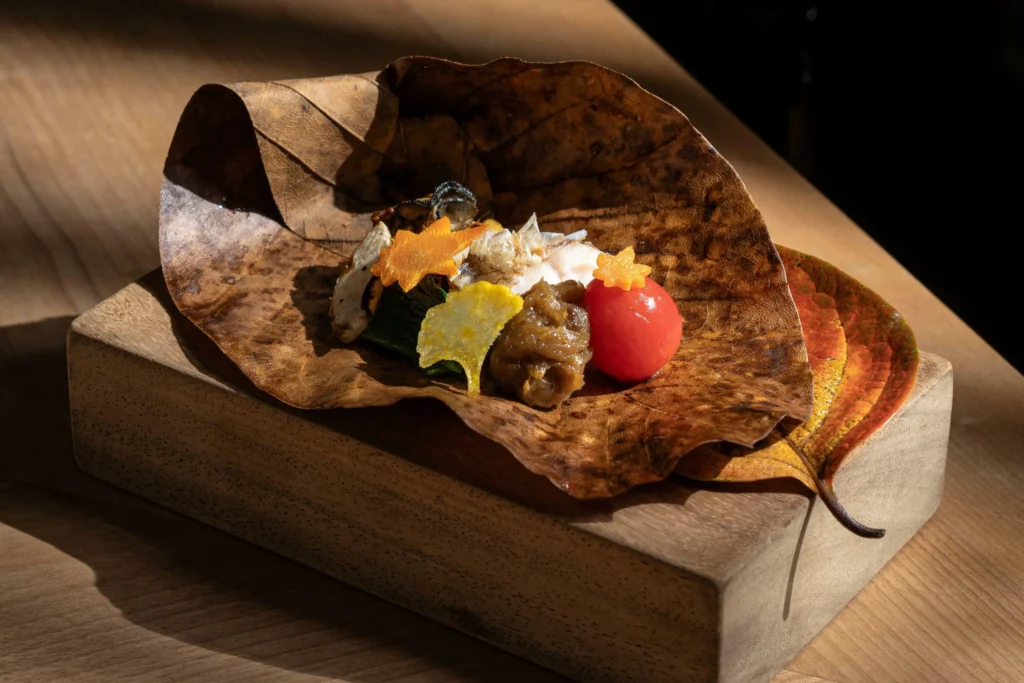 An elegant Japanese dish is beautifully arranged on a large, dried brown leaf resting atop a rectangular wooden block. The presentation features small, delicate portions of fish, greens, paste, and a bright red cherry tomato, accented by vibrant, autumn-themed garnishes shaped like stars and leaves.