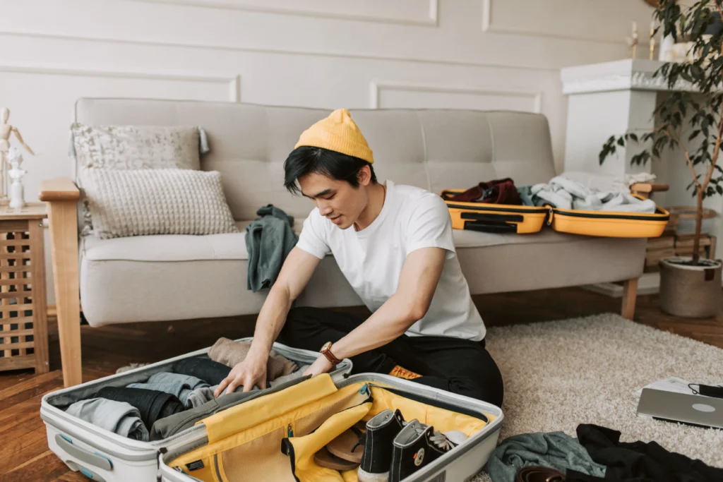 A young man wearing a white t-shirt and a bright yellow beanie sits on the floor of a living room, carefully packing clothes into a large grey suitcase. In the background, an open yellow suitcase rests on a beige couch surrounded by loose clothing and travel gear.