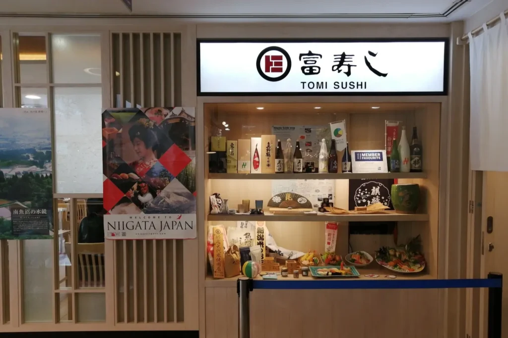 The image features the illuminated storefront sign of "TOMI SUSHI" set above a wooden display case filled with sake bottles, food models, and decorative Japanese items. To the left, a large colorful poster welcomes visitors to Niigata, Japan alongside glass windows with light wood framing.
