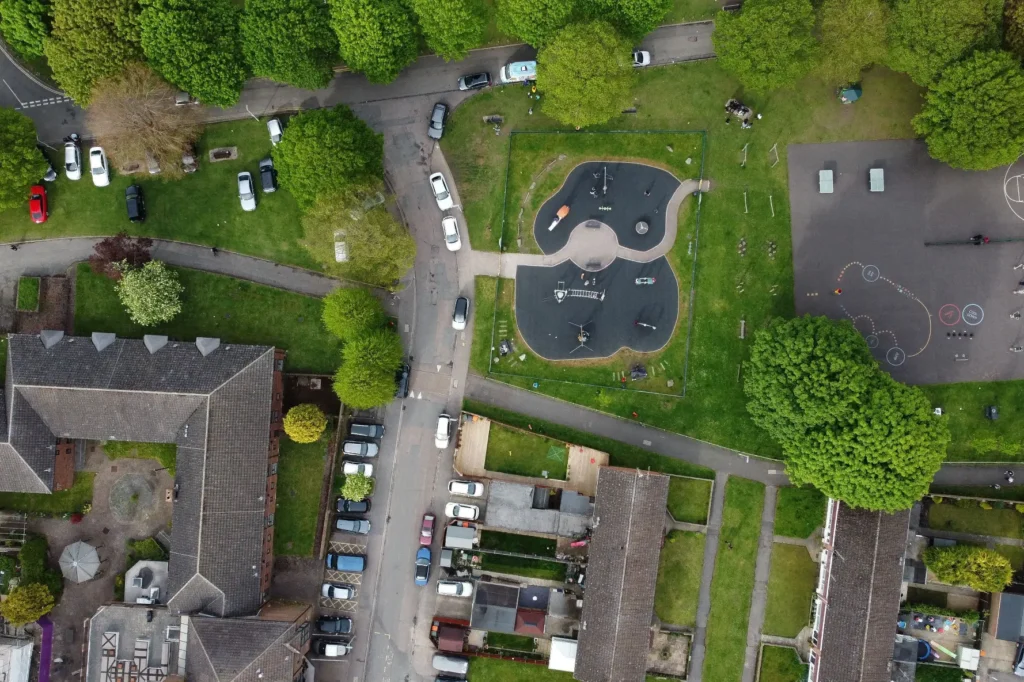 An aerial, top-down view shows a neighborhood park featuring a modern playground with a dark safety surface shaped like a figure-eight. Surrounding the park are lush green lawns, mature trees, residential buildings with tiled roofs, and streets lined with parked cars.