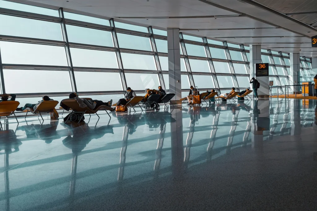Travelers relax and nap on lounge chairs inside a spacious airport terminal in front of massive, angled floor-to-ceiling windows. Bright natural light reflects off the polished floor, casting long shadows and grid patterns throughout the waiting area near gate 36.