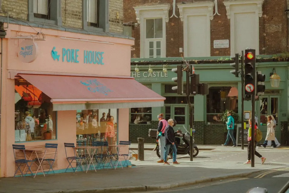 A street view of a pastel pink restaurant named "Poke House" featuring outdoor seating, large windows, and a matching pink awning on a city corner. In the background, pedestrians walk past a green-fronted pub called "The Castle" on Portobello Road near a traffic light.