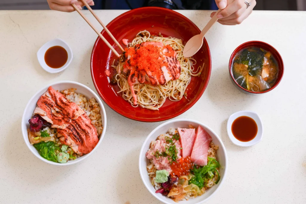 A top-down view shows a meal spread featuring Japanese dishes, including fresh and seared seafood rice bowls alongside a side of miso soup. In the center, a person uses chopsticks and a wooden spoon to eat from a large red bowl filled with thin noodles topped with creamy seared sauce and bright orange fish roe.