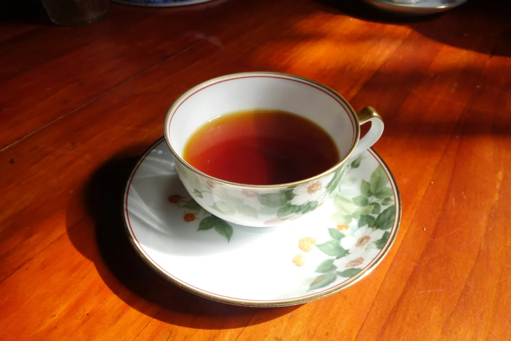 A porcelain teacup and matching saucer adorned with green leaf and white flower patterns sit on a polished wooden table. Sunlight streams across the surface, casting a soft shadow beside the cup of dark, amber-colored tea.