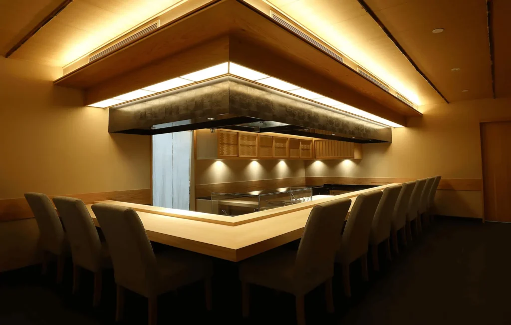 An elegant, modern Japanese dining room featuring a sleek L-shaped wooden counter surrounded by comfortable upholstered high-back chairs. The space is illuminated by warm, indirect cove lighting along the wooden ceiling and drop-down canopy, creating an intimate and sophisticated atmosphere.