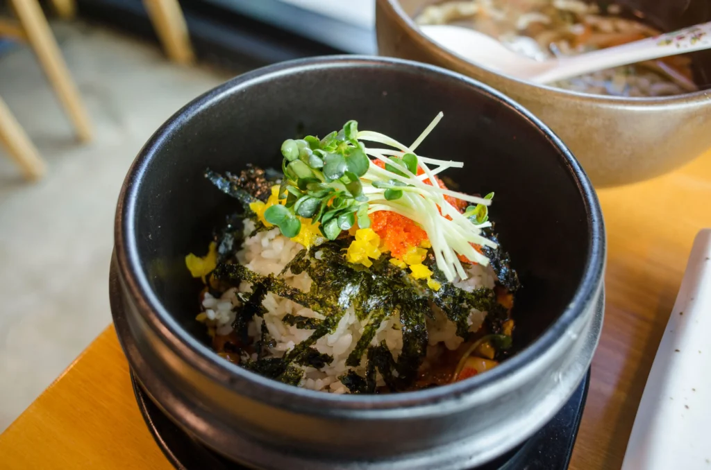 A dark stone bowl filled with white rice is artfully topped with shredded seaweed, yellow pickled radish, bright orange fish roe, and microgreens. The dish sits on a wooden restaurant table with a bowl of soup visible in the background.