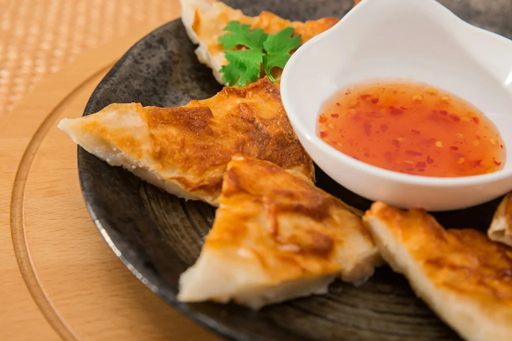 Several golden-brown, crispy triangular pastries or pancakes are arranged on a dark plate, garnished with a single sprig of cilantro. A small white bowl filled with a red, flecked sweet chili dipping sauce sits alongside them on a round wooden board.