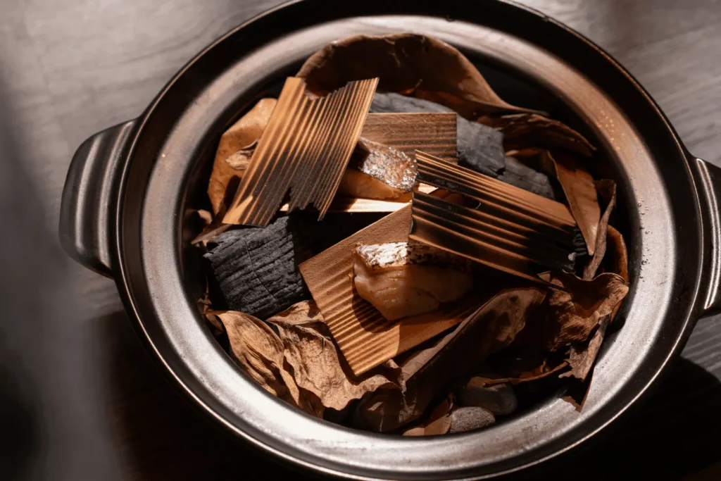 A top-down view of a dark ceramic cooking pot containing pieces of fish presented on thin, ribbed wood planks. The fish and planks are artistically arranged inside the pot over a rustic bed of dried leaves, charcoal chunks, and small stones.