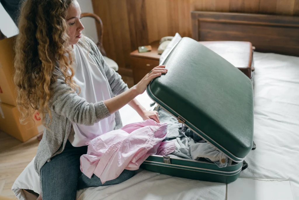 A young woman with long curly blonde hair sits on a bed, carefully packing clothes into a large, open green vintage suitcase. She holds a light pink shirt in her hands while arranging items inside the luggage, surrounded by cardboard moving boxes and a brown wooden headboard in the background.